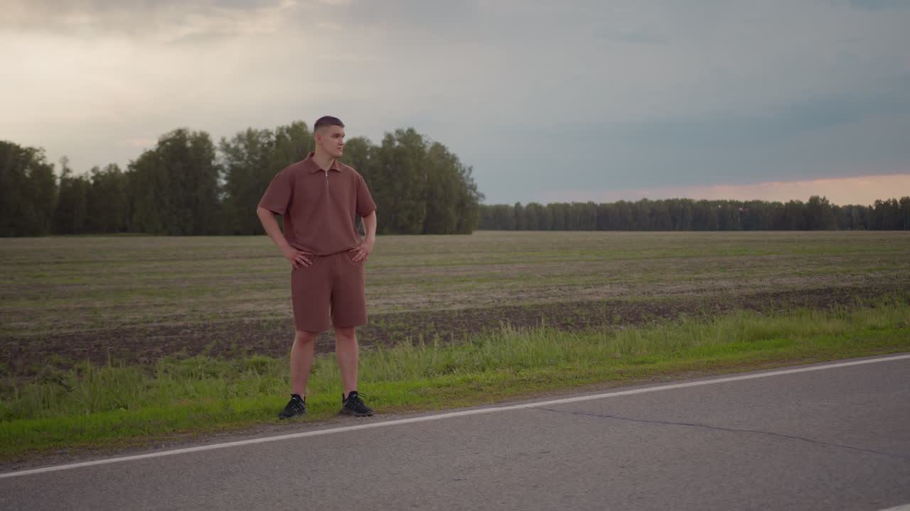 Young man in brown casual outfit stands confidently by roadside with hands on waist, gazing into distance over plowed field under moody sky, conveying calm, awareness, and solitude in rural setting