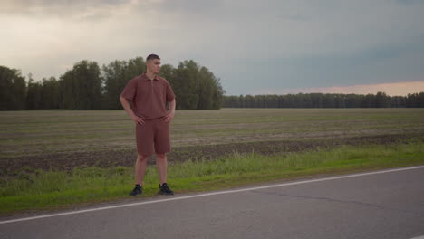 young man in brown casual outfit stands confidently by roadside with hands on waist, gazing into distance over plowed field under moody sky, conveying calm, awareness, and solitude in rural setting