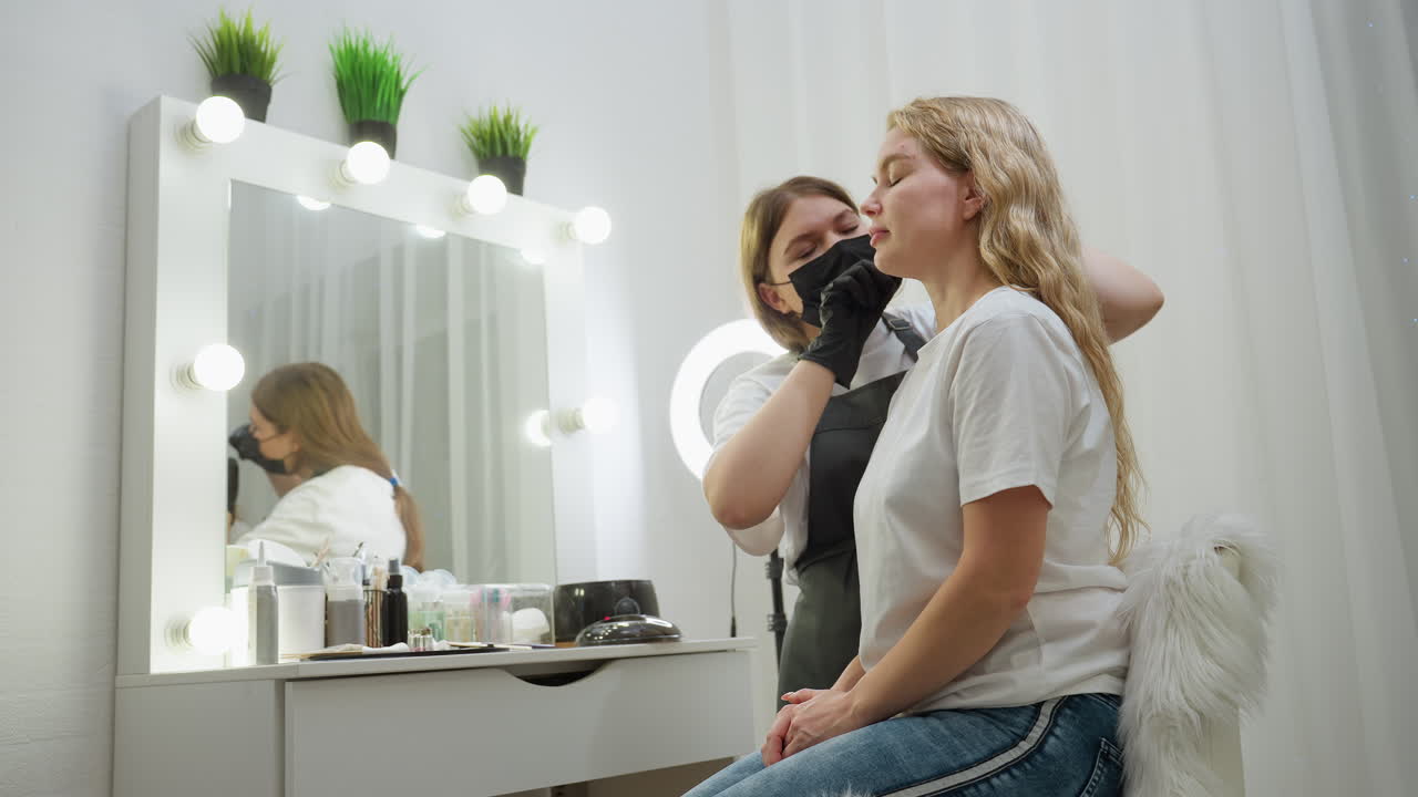 Beautician wearing white top and black gloves concentrates while attending to client seated on fluffy chair. Background features beauty tools, illuminated mirror, and decorative green indoor plants