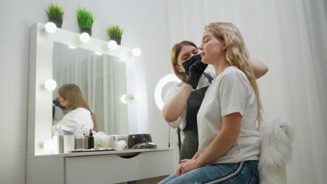 beautician wearing white top and black gloves concentrates while attending to client seated on fluffy chair. background features beauty tools, illuminated mirror, and decorative green indoor plants