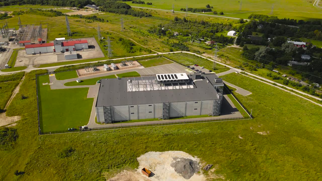 Aerial drone footage of a large modern data center and a power distribution electrical station next to it at sunny summer countryside in the Baltics. The server room has cooling system fans on rooftop