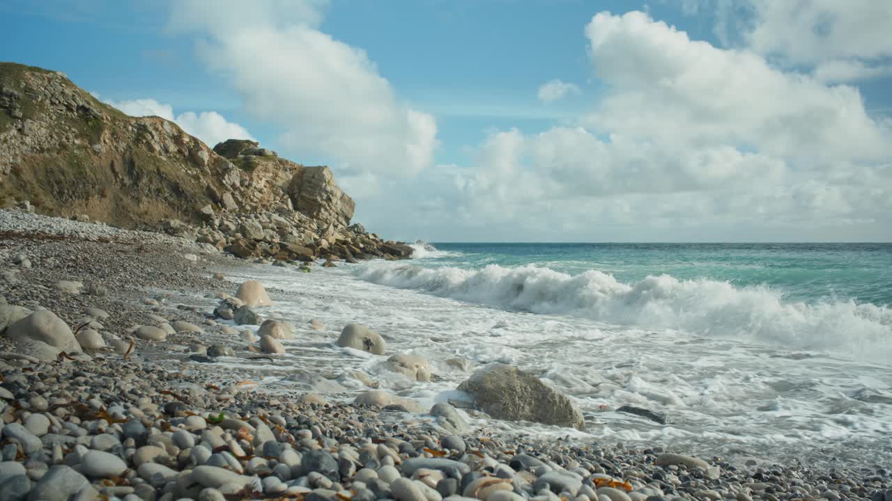 Scatto panoramico cinematografico in 4K di onde che si infrangono su una spiaggia rocciosa in una giornata di sole, a Church Ope, sull'isola di Portland, nel Dorset, Inghilterra