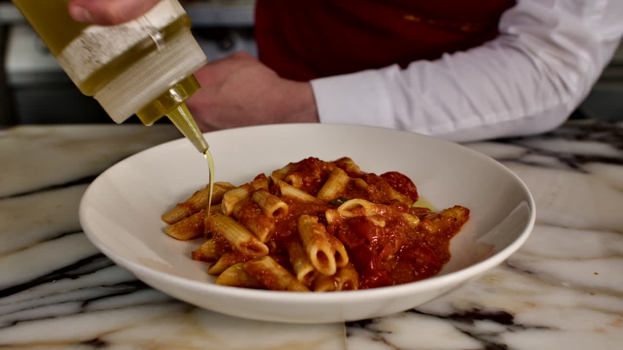 Primo piano di uno chef che irrora olio extra vergine d'oliva su un piatto di penne al sugo di pomodoro in una cucina generica. Food styling professionale e tocco finale