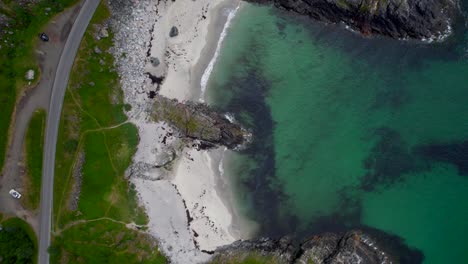 Aerial-birdseye-view-of-beach-in-Andoya-Norway-in-summer-with-green-water-and-grass-along-a-rocky-show-and-curving-road
