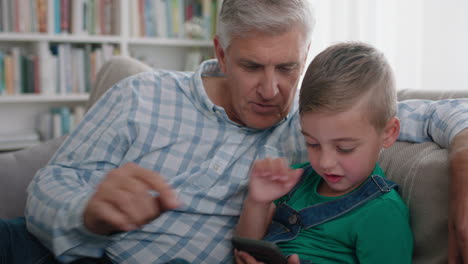Glücklicher-Großvater,-Der-Dem-Kleinen-Jungen-Zeigt,-Wie-Man-Das-Smartphone-Benutzt,-Und-Dem-Neugierigen-Enkel-Moderne-Technologie-Beibringt,-Intelligentes-Kind,-Das-Das-Mobiltelefon-Lernt,-Während-Es-Mit-Dem-Opa-Auf-Dem-Sofa-Sitzt