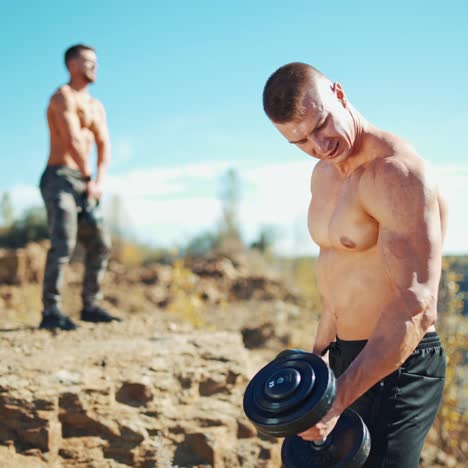 sportsmen doing workout outdoors. muscular athlete is lifting dumbbells in hands on the background of a shirtless man with kettlebell in summer.