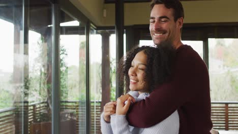 Happy-diverse-couple-embracing-and-laughing-in-living-room,-looking-out-of-window-in-the-countryside