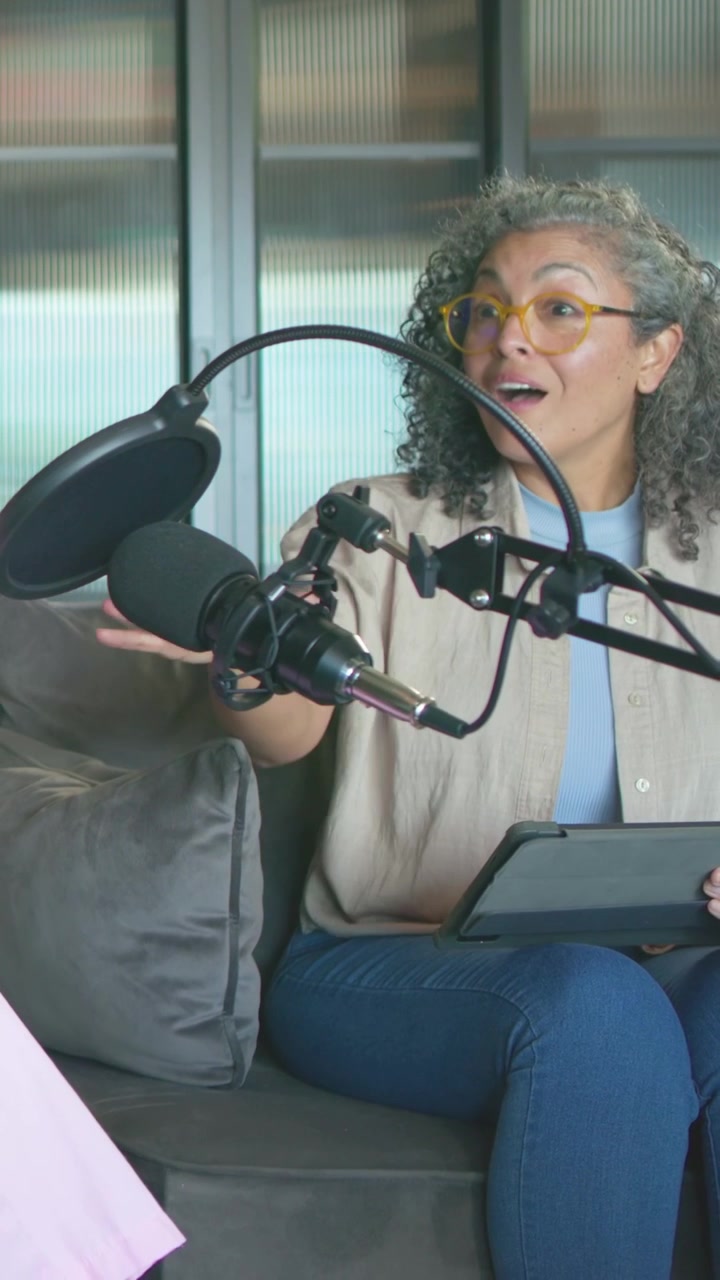 Mujer mayor afroamericana con gafas amarillas hablando por un micrófono y leyendo una tablet en un sofá en un estudio.