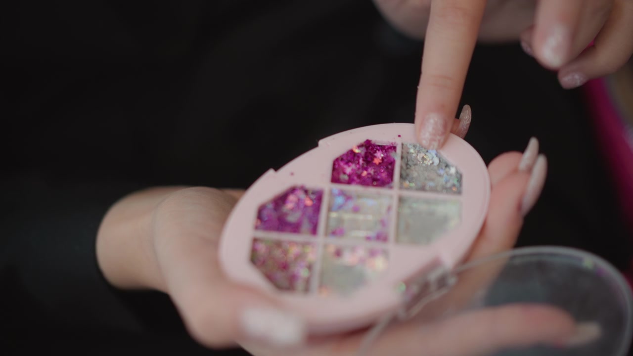 Close macro view of hands handling circular compartmented nail art organizer while another hand pinches metallic glitter and places it into small sections, showing careful beauty preparation