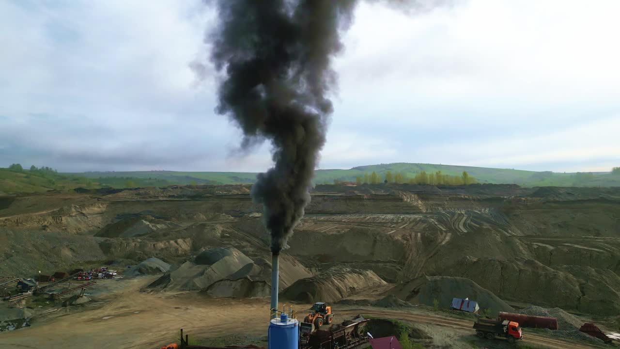 Fumo nero si innalza da un'alta ciminiera industriale. Colline di terra e macchinari circondano la fabbrica fumosa. Gli operai azionano le macchine in un'ampia cava a cielo aperto. Il fumo si innalza contro un cielo nuvoloso.