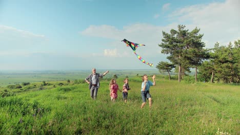 caucasian senior man running with his grandchildren in the park while they are flying a kite on a sunny day