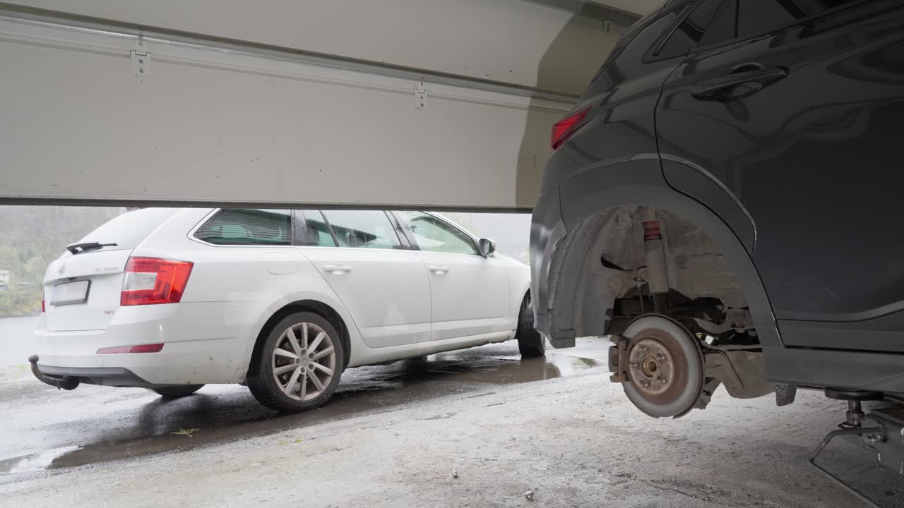 Garage door closing with small gray car inside on lift, rear wheel removed, white car outside