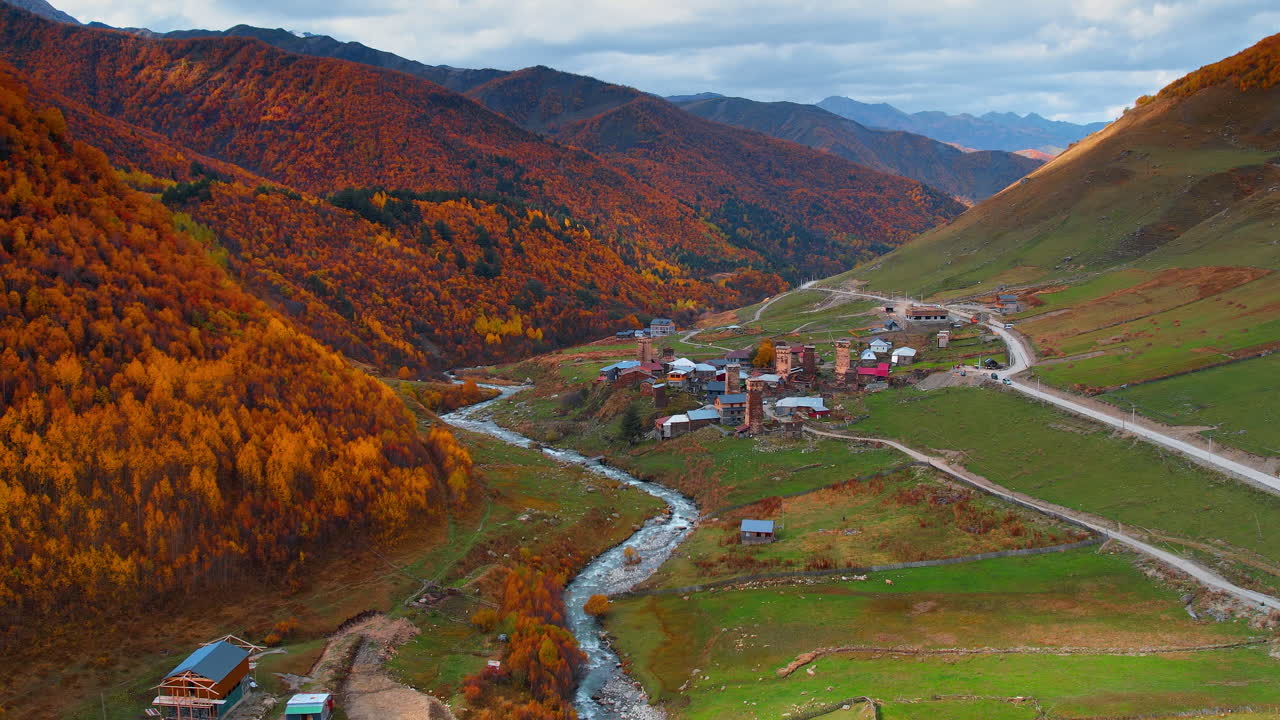 Voando sobre o rio sinuoso que flui entre as rochas. Aproximando-se da área residencial com casas-torre medievais na vila de Ushguli, Alto Svaneti, Geórgia. Florestas de um laranja vibrante cobrem as montanhas no outono.