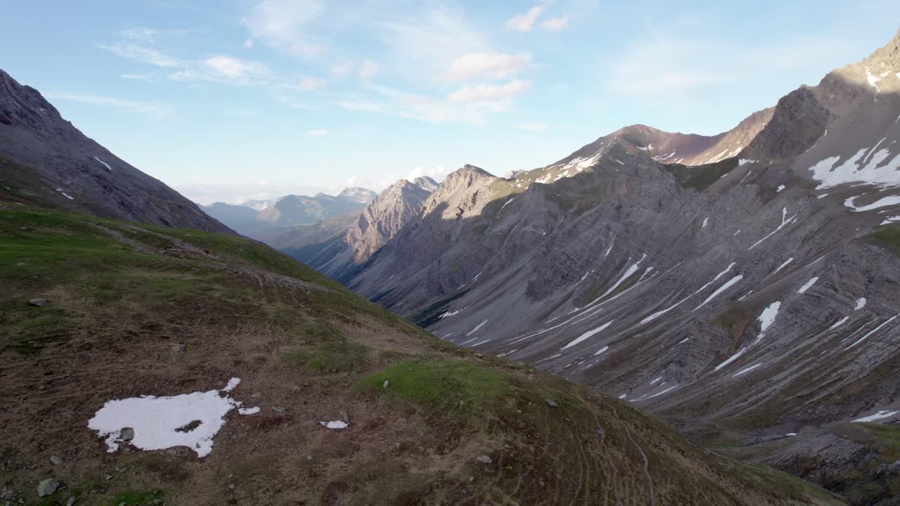 Aerial drone footage slowly rising upwards and looking across a dramatic, jagged mountain landscape with residual patches of snow and alpine meadows looking down a glacial valley in Switzerland