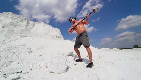 strong man with steel hammer works on white hill under blue sky. sporty male in shorts breaks big stone by heavy hammer outdoors.