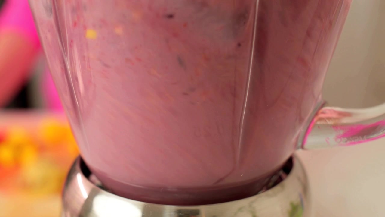 Primer plano extremo de frutos rojos, mango y leche girando dentro de una licuadora de vidrio y transformándose en un batido de frutas cremoso y rosado en una luminosa cocina casera.