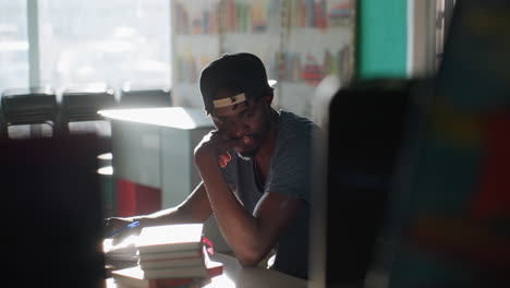 young man wearing cap sits in library studying at table with stacks of books, resting chin on hand while looking thoughtful, teal and orange walls and shelves behind, warm light creating calm mood