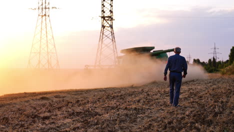 Rear view of a man walking by the field of mowed wheat. A modern combine harvester works in the cloud of smoke at backdrop