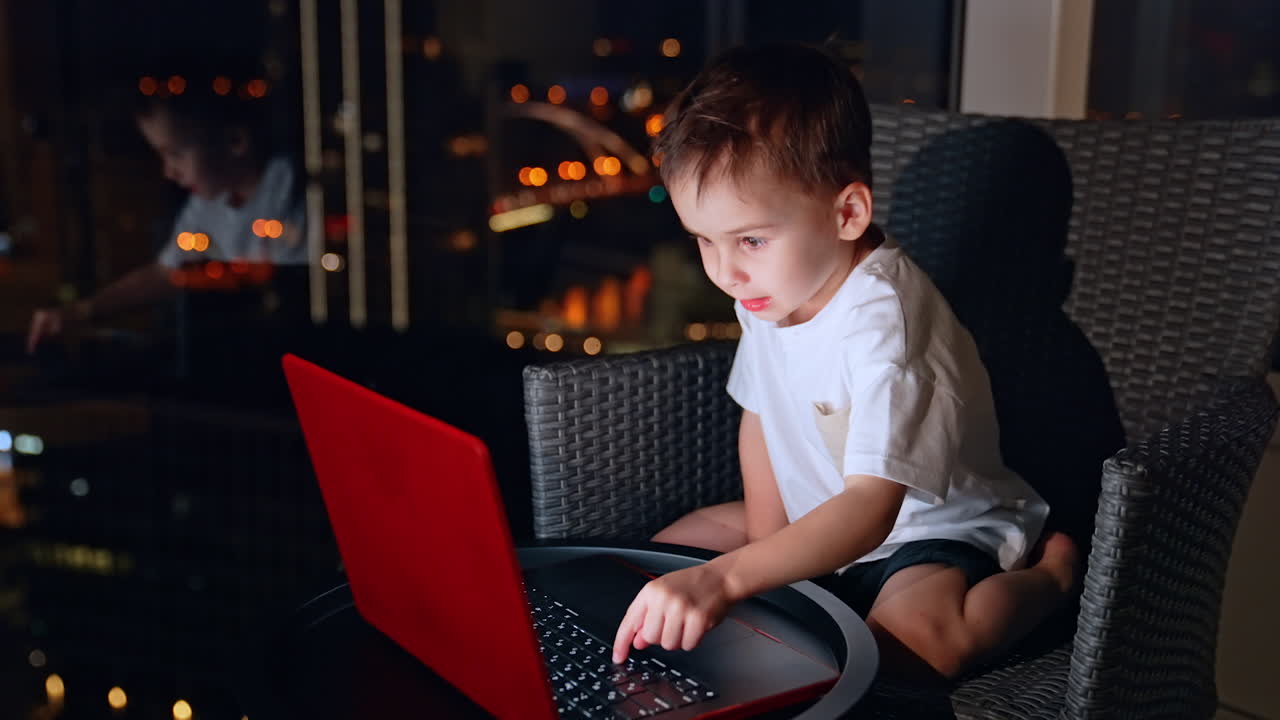 Child smiling while using laptop on balcony at night. A young boy smiles while using a laptop on the balcony at night, with city lights glowing in the background