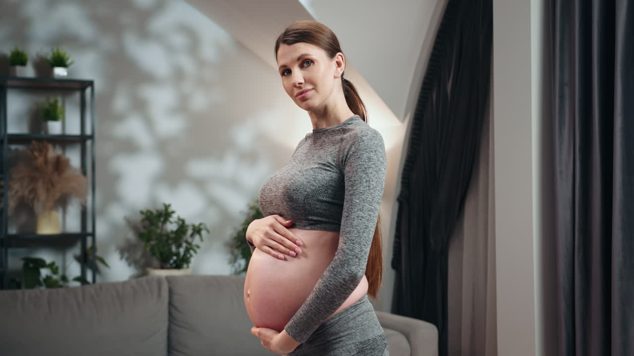 Pregnant woman in athletic wear cradles her belly standing in a softly lit room with home decor
