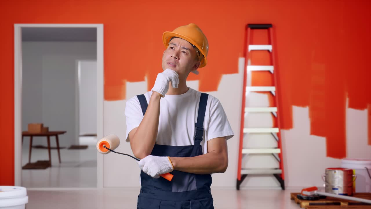 Asian Man Painter Wearing Safety Helmet Thinking About Something And Looking Around While Standing With Painting Wall In Room