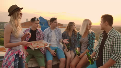 the company of six young people communicate with each other on the roof with pizza and beer. girls look around at summer sunset