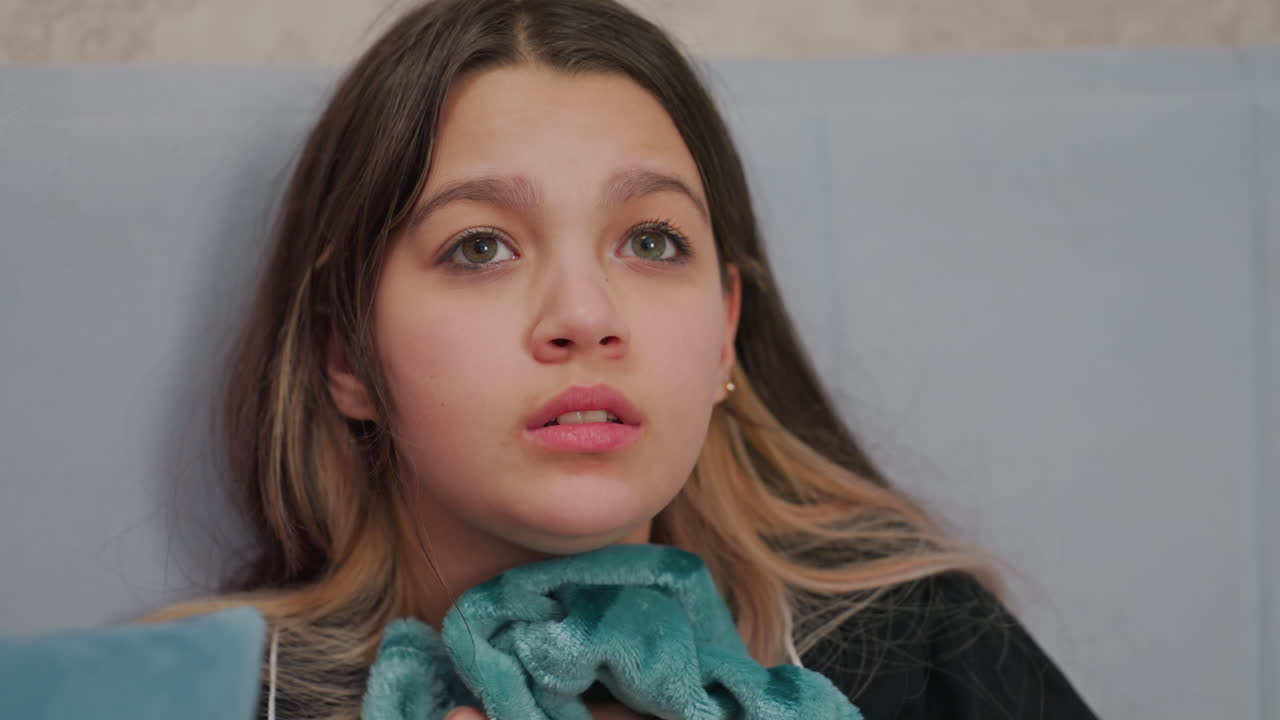 close up of young girl sitting against soft headboard gripping fuzzy blanket while watching intense scene in movie, eyes partially shut and lips pressed in nervous expression