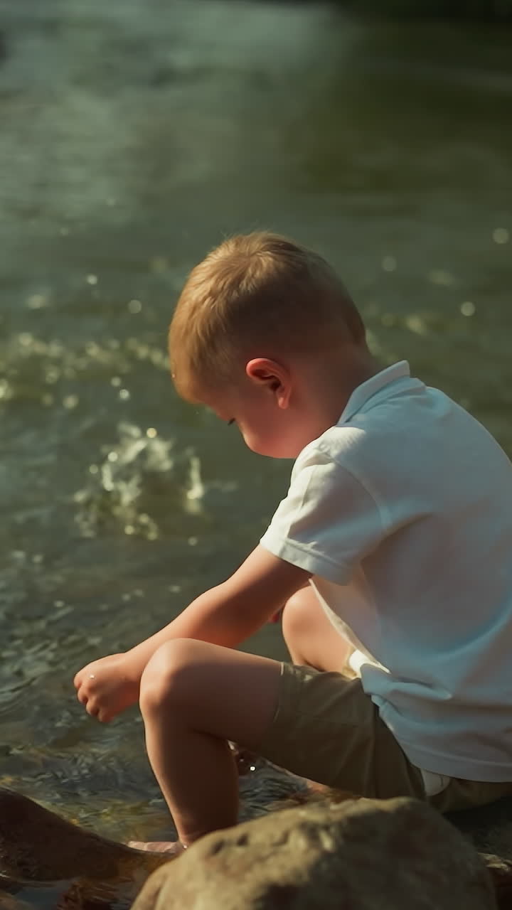 Young boy plays in park throwing stones into shallow river water at sunset. Little child relishes atmospheric evening with amazing landscape and clear water