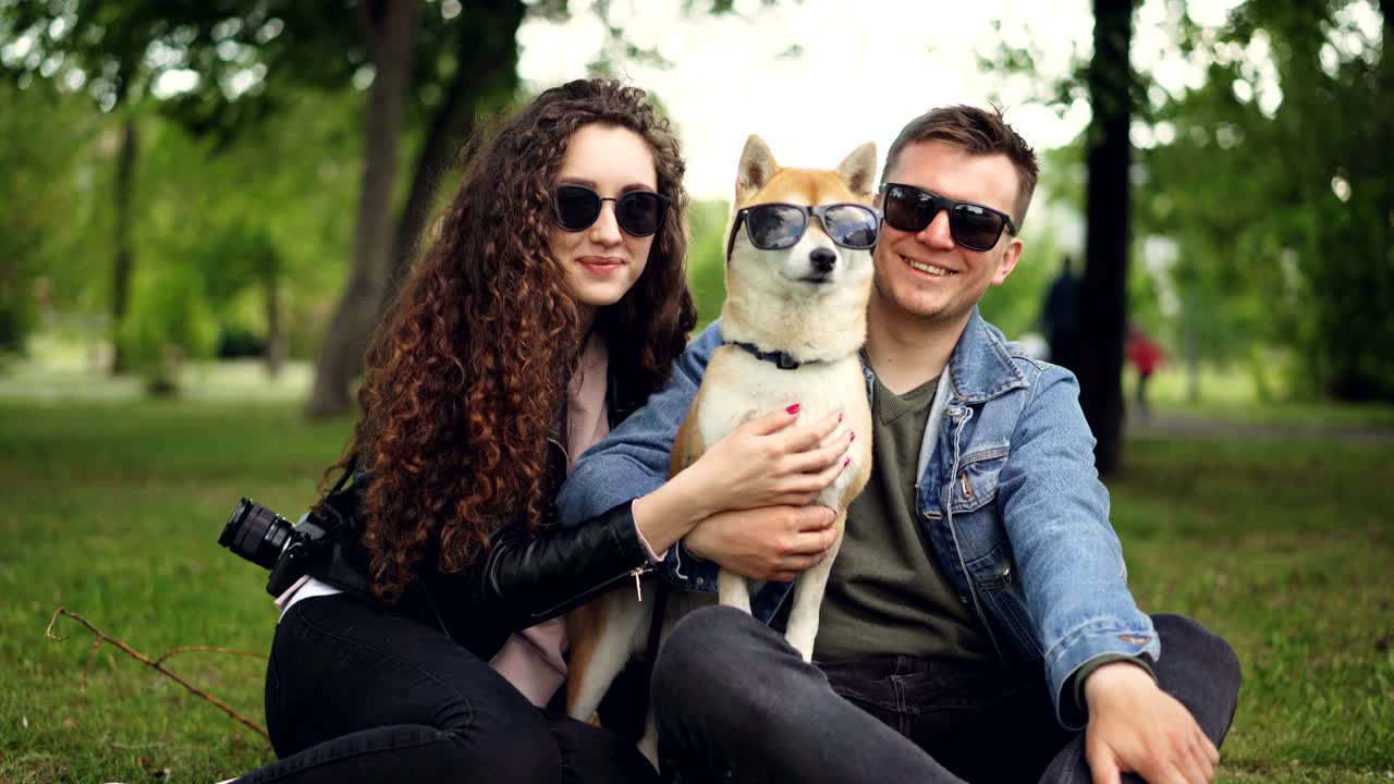 retrato de una pareja linda y un perro shiba inu todos con gafas de sol mirando a la cámara y sonriendo sentados en la hierba en el parque. estilo de vida moderno, animales lindos y concepto de naturaleza.