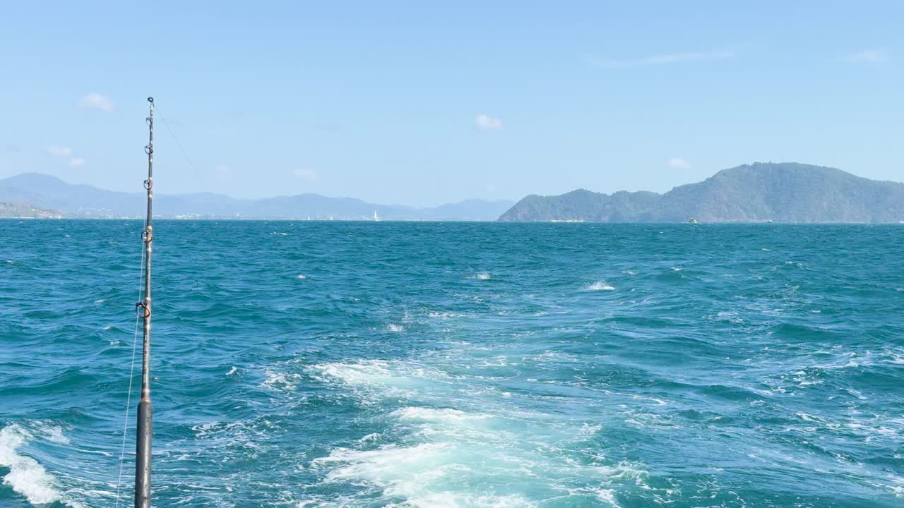 A boat creates a wake in the ocean near Phuket, Thailand, under clear blue skies with distant islands visible