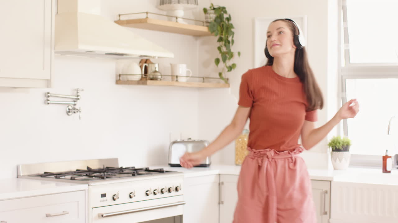 Giovane donna in cucina che balla con gioia mentre ascolta musica con le cuffie