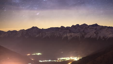Zeitraffer-Von-Sternen,-Die-Sich-Am-Himmel-über-Schneebedeckten-Felsigen-Bergen-Mit-Beleuchtetem-Dorf-Im-Tal-Bewegen
