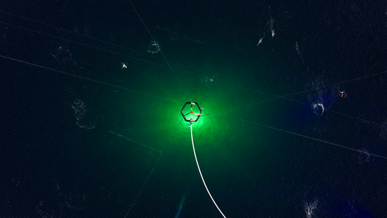 Illuminated salmon farm cage and net structure in dark Arctic waters, aerial top down overview