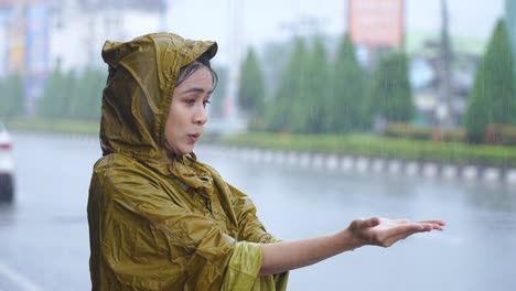 young tired female office worker in yellow raincoat stuck in a rain, stands under roof and playing with hands collecting raindrops, heavy rain caused by monsoon, asian city life, tropical zone country