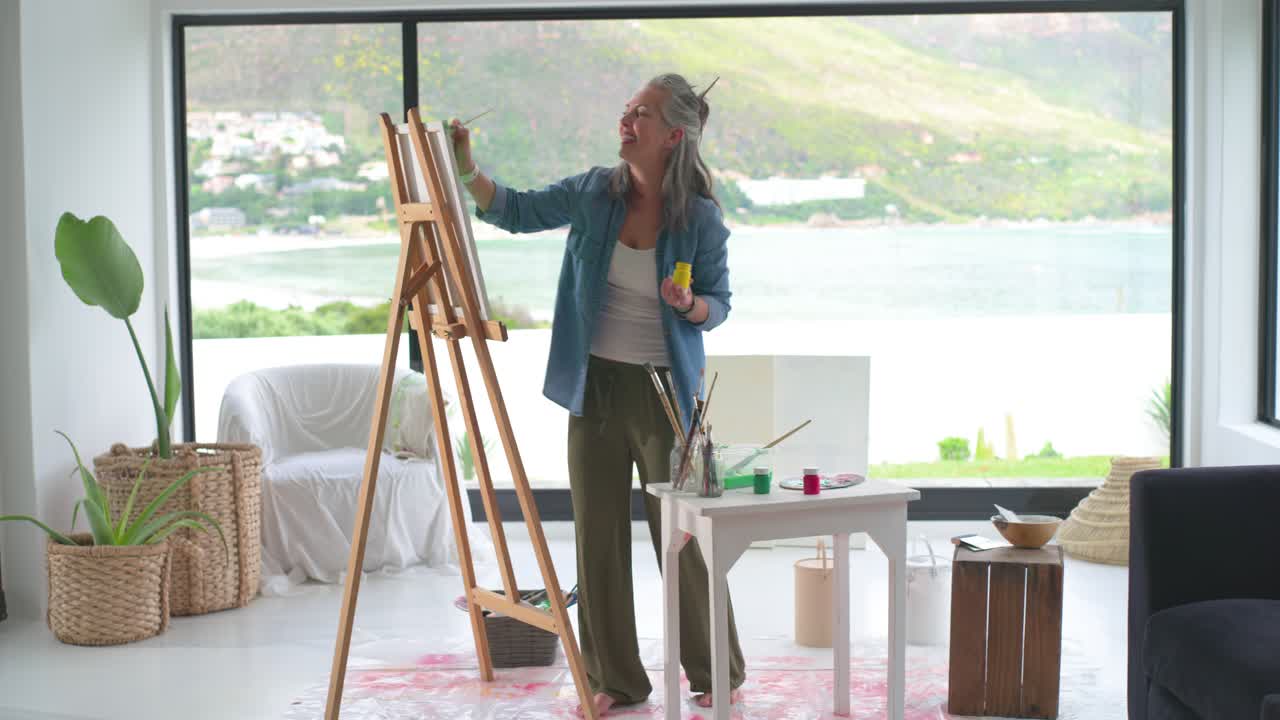 Senior female painting on easel in home studio, selecting yellow paint and brushes, creating art