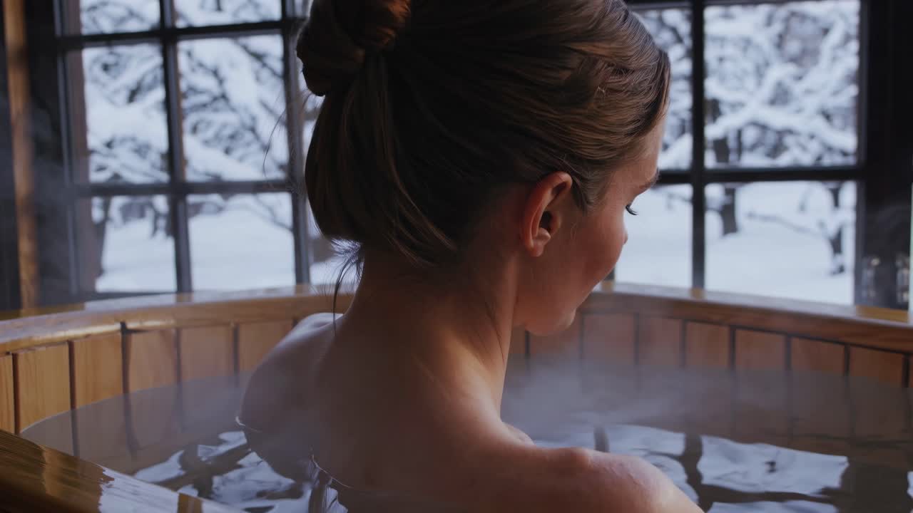 A serene video scene from behind, capturing a woman in a wooden hot tub, gazing out at a snowy