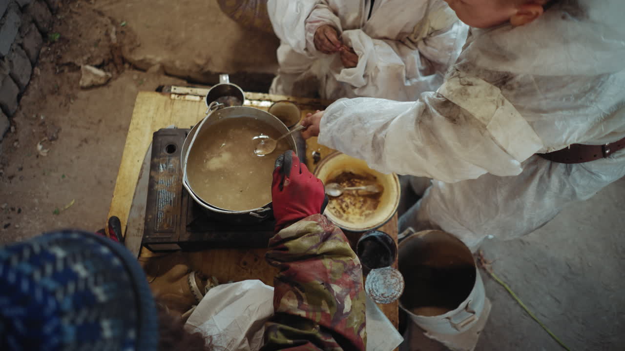 Overhead view shows survivors in worn protective clothing preparing food in rusty pot on old stove inside abandoned shelter, stirring simple meal surrounded by dirty utensils and orphaned children