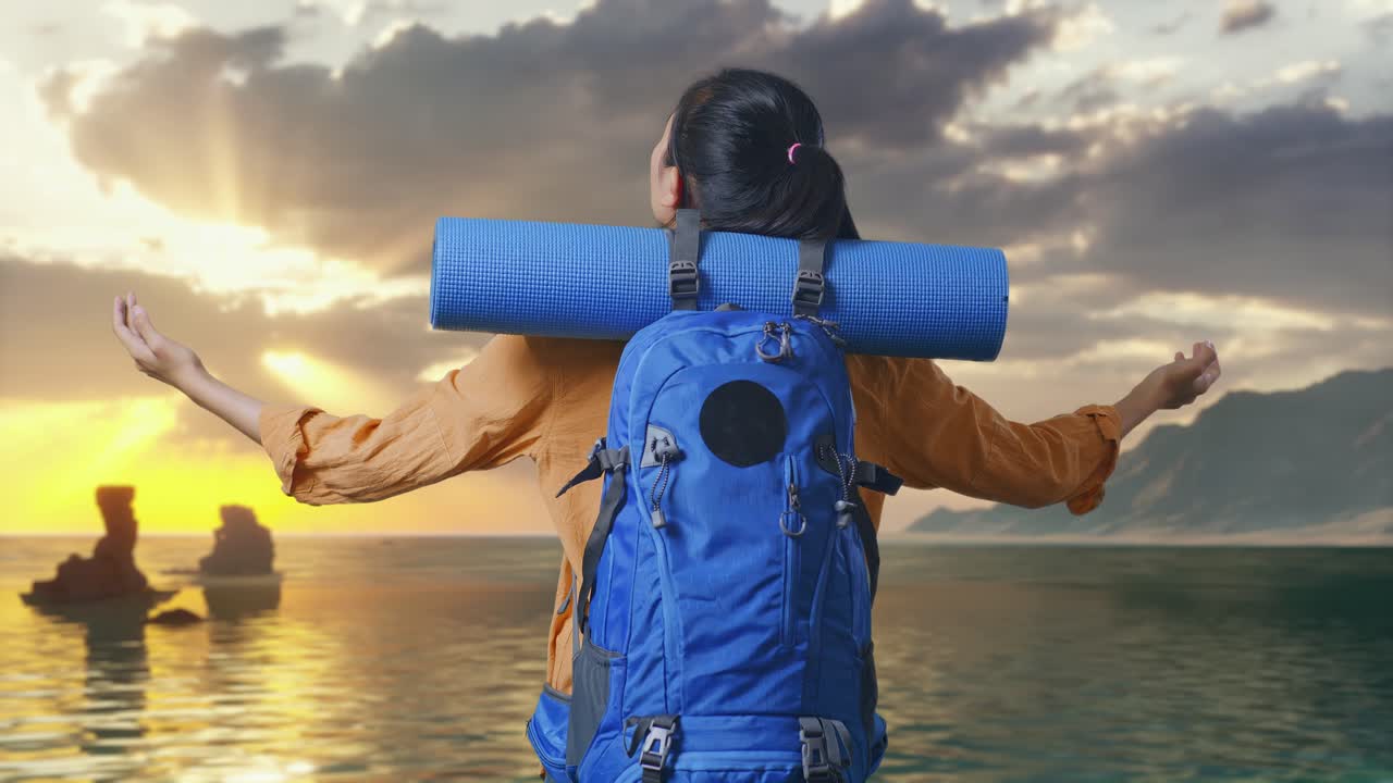 Vista posterior de una senderista con mochila de alpinismo abriendo los brazos y observando el paisaje mientras está de pie en la playa al atardecer