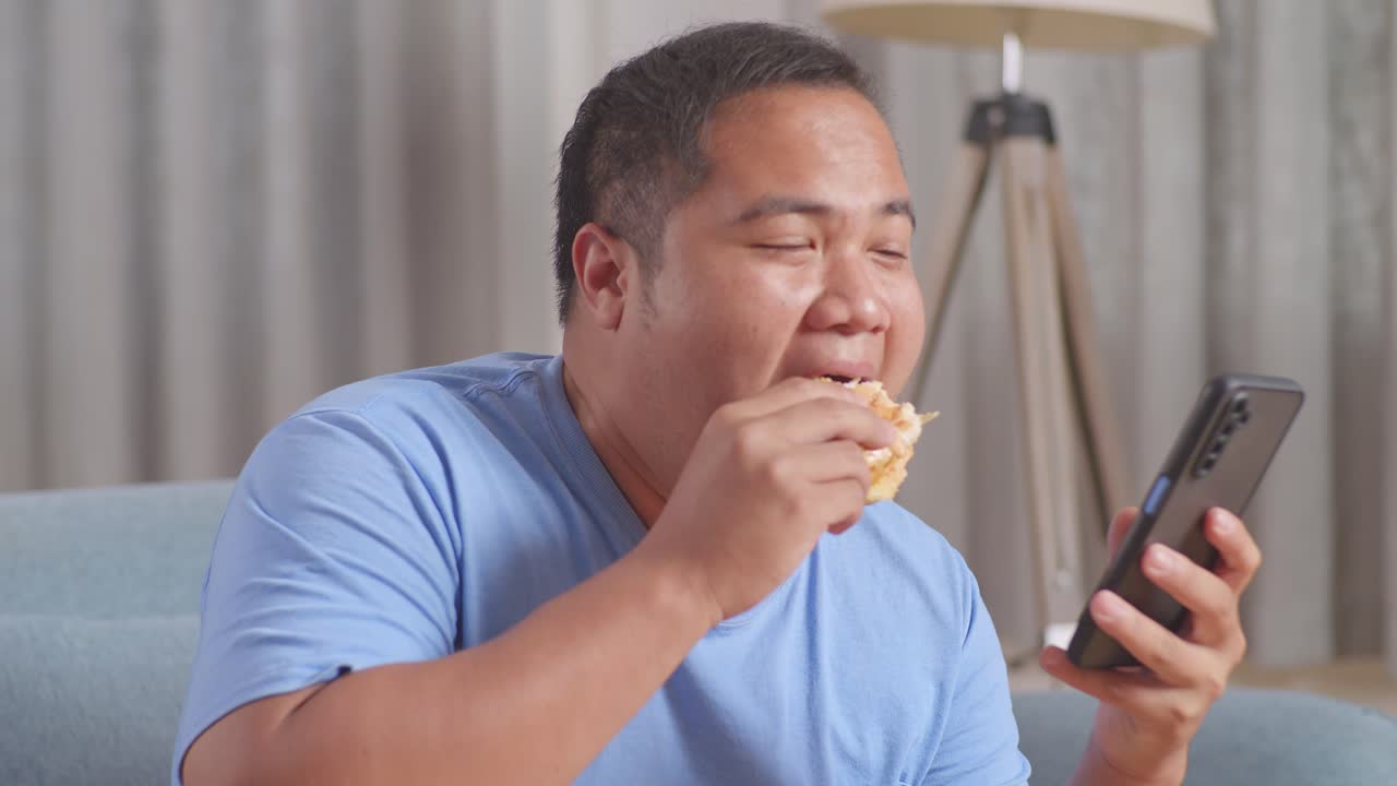 한 아시아 남자가 집에서 거실에서 소파에 긴 을 먹고 패스트푸드를 먹는 동안 스마트폰을 연주하고 있습니다.