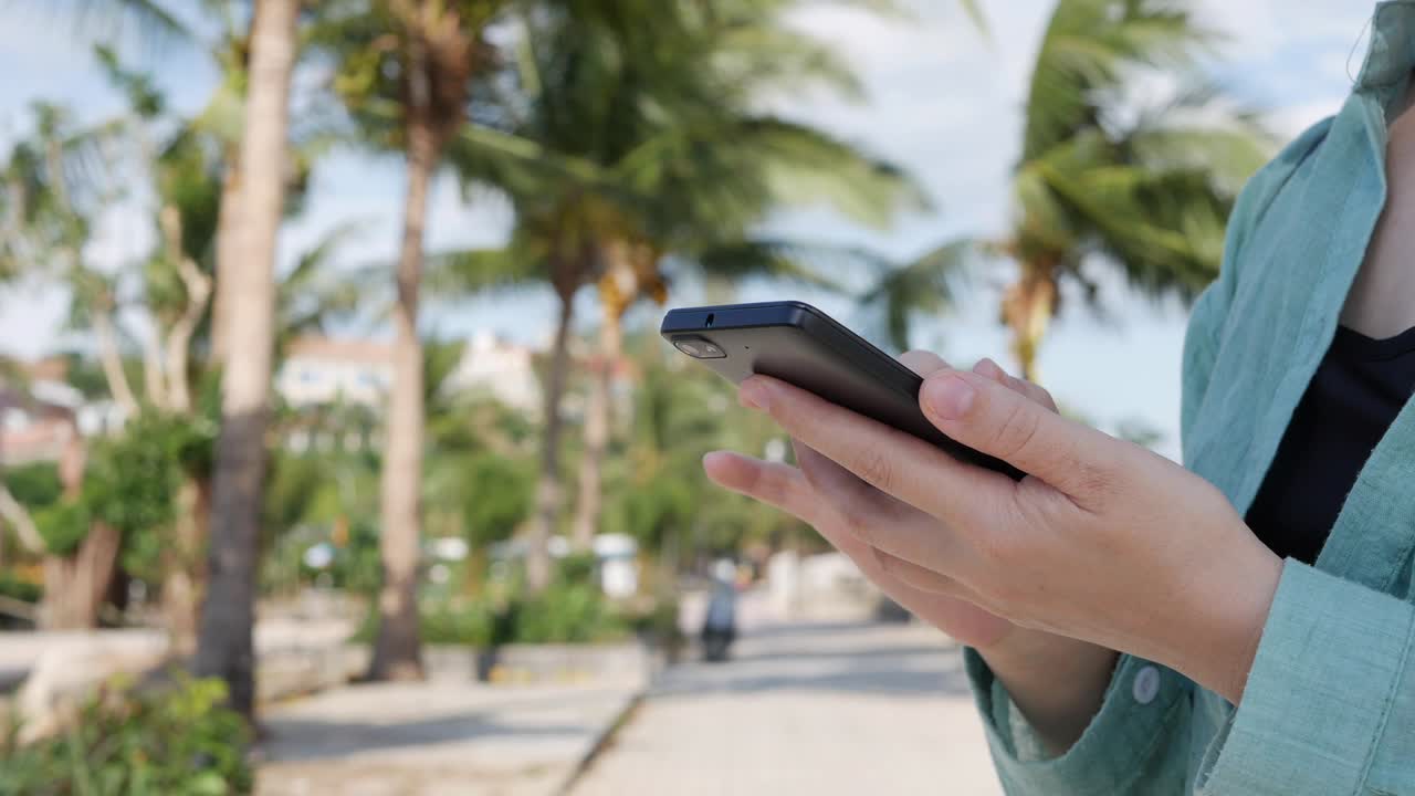 hermosa mujer delgada con el cabello largo rubio en camisa verde de pie cerca de la palmera y el uso de teléfono inteligente sobre el fondo del parque. niña en la pantalla cuadrada tocando y sonriendo