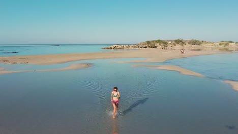 backwards drone aerial view woman running in empty elafonissi beach lagoon reflection of blue clear sky