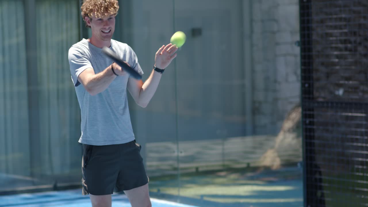 Mann i 20-årene skifter vekt, kaster en neonball og svinger en svart racket på en blå bane mens han øver på serve.