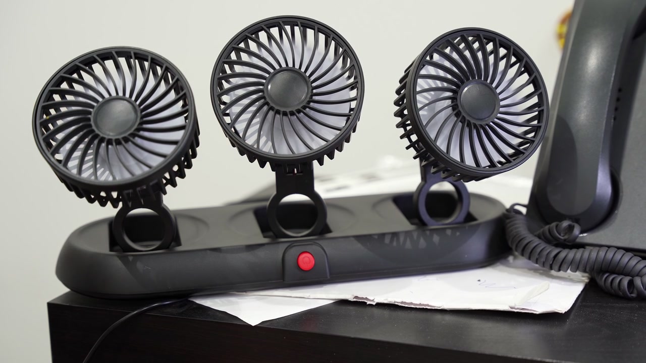 Nærbillede af mandehænder, der samler en sort, trehovedet bærbar ventilator på et mørkt skrivebord indendørs. En fastnettelefon står i nærheden i et lyst, moderne kontorarbejdsområde, klar til sommervarmen.