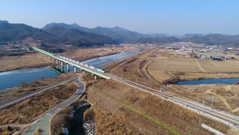 High-speed-train-passing-from-a-bridge-over-a-river-with-blue-water-and-mountains-in-the-background