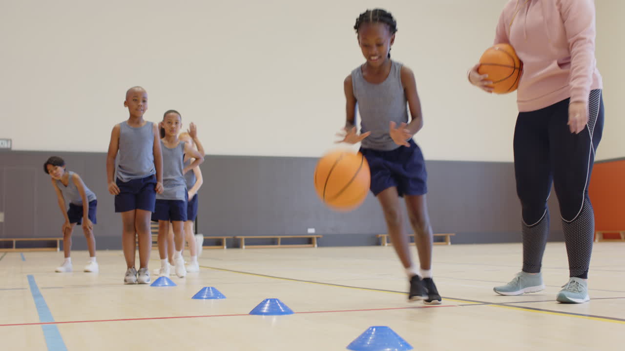 Dribbling basketball around cones, children practicing in school gym with coach