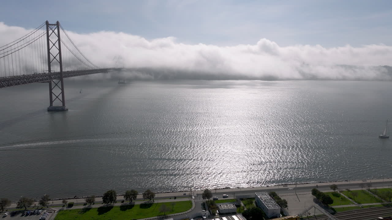 Aerial drone shot towards the 25th April Bridge and waterfront in Lisbon, Portugal, Europe. Sunny and bright with clouds, fog, mist covering the bridge