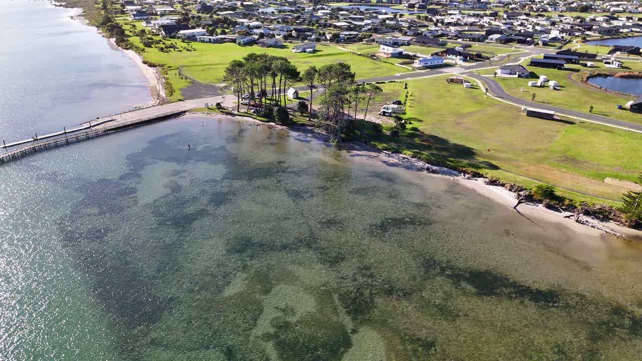 En 4K-drone svæver over stranden og molen ved Matarangi Freedom Camp i New Zealand. Sandstrande, roligt søvand og frodige omgivelser skaber en fredfyldt og smuk udsigt.