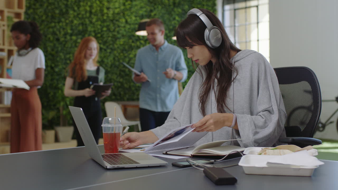 joven mujer de negocios asiática escribiendo notas disfrutando del estudio escuchando música usando auriculares estudiante trabajando en una oficina multiétnica ocupada