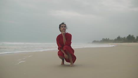 a beautiful young indian girl with a beautiful red dress is sitting on the seashore and is looking at the waves on a cloudy day