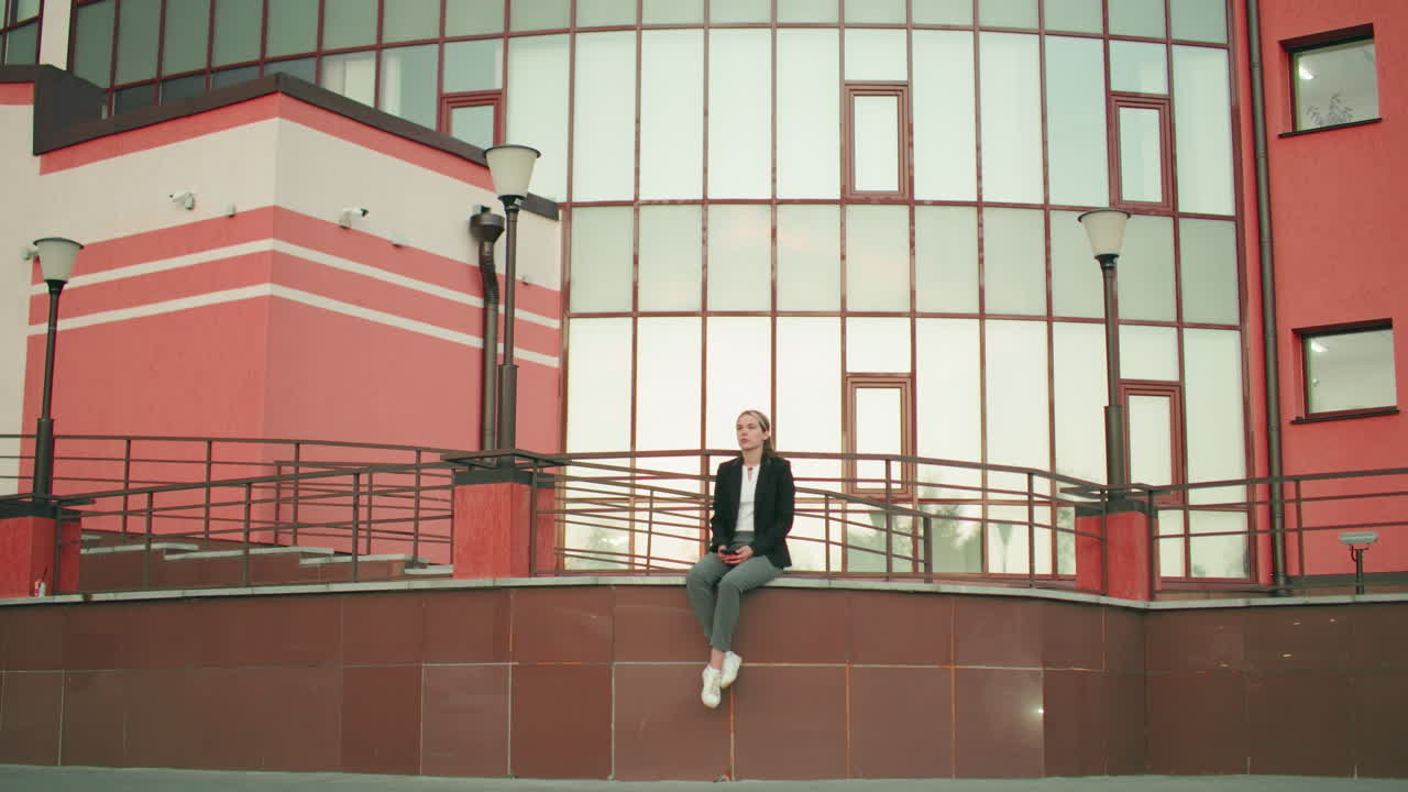 Sober lady in black blazer sits alone on fence with legs crossed at ankle holding phone, looking thoughtful and calm in urban setting with modern building and lampposts in soft daylight background